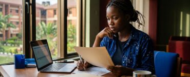 Nigerian student reviews admission papers beside a laptop and folder near a campus window.