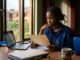 Nigerian student reviews admission papers beside a laptop and folder near a campus window.