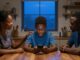 A Nigerian teenager uses a phone while his parents sit beside him in a warm kitchen.
